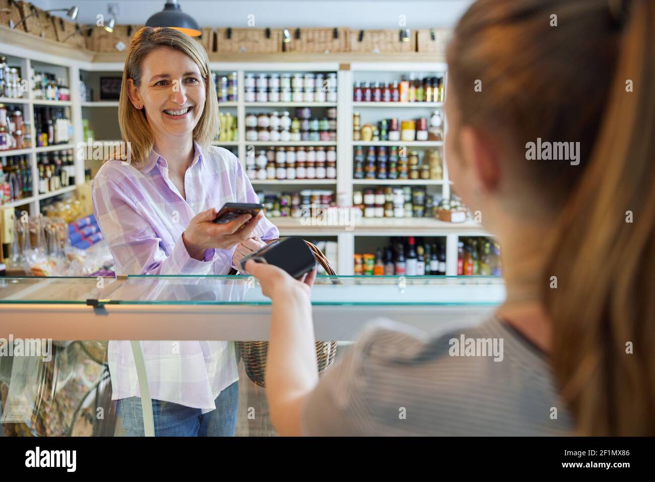 Smiling Female Customer Delicatessen Food Store Making Contactless Payment With Mobile Phone For Shopping To Sales Assistant Stock Photo