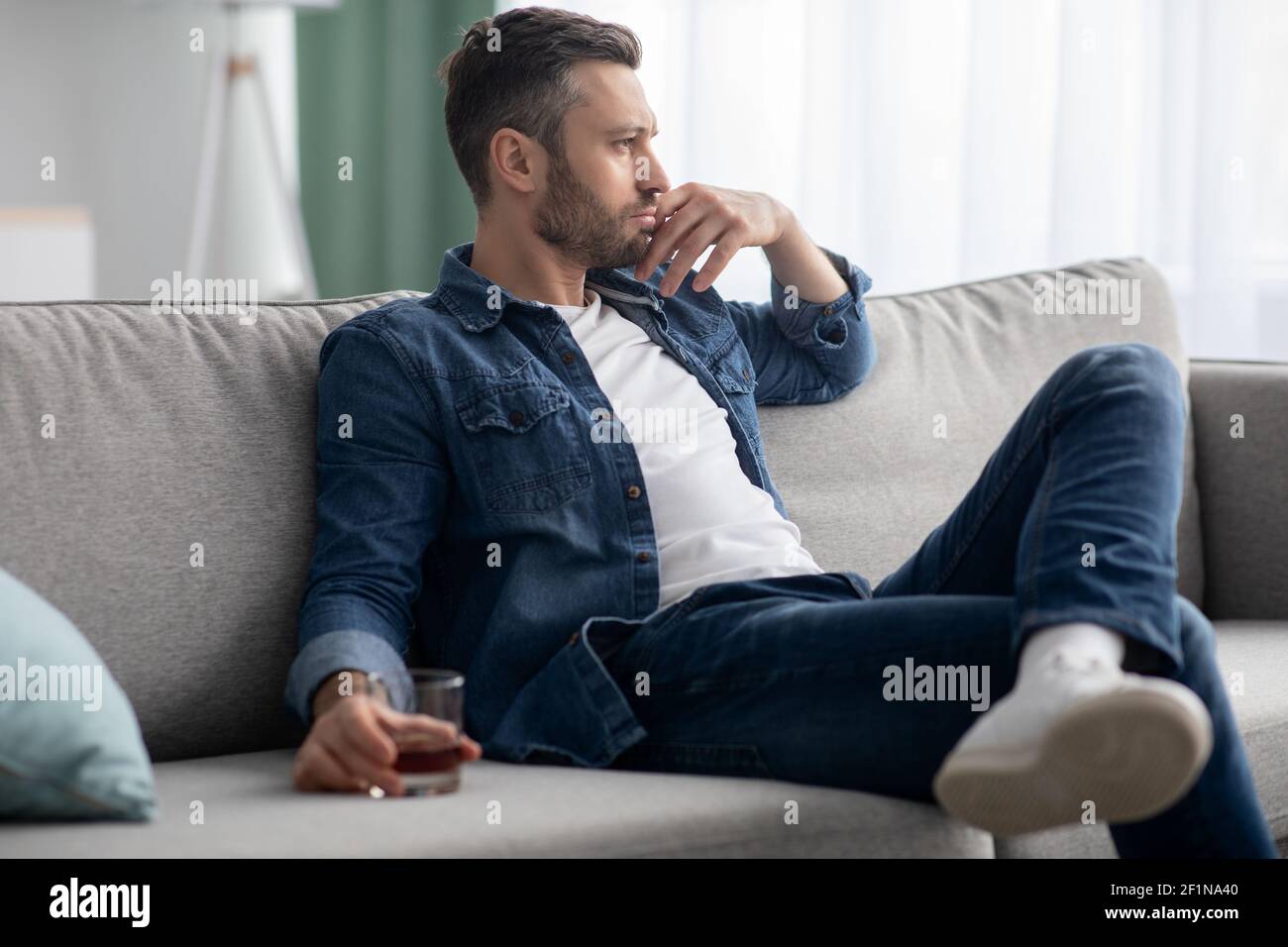 Depression, crisis concept. Sad middle-aged bearded man sitting on sofa, drinking alcohol at home, feeling lonely and sick, holding glass of whiskey a Stock Photo