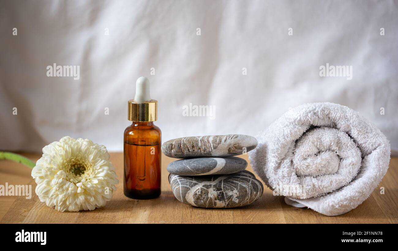 Dark glass vessel with essential oil cosmetic product for skin care, massage and spa at home. Stack of stones, hand towel and white flower on the wood. Stock Photo