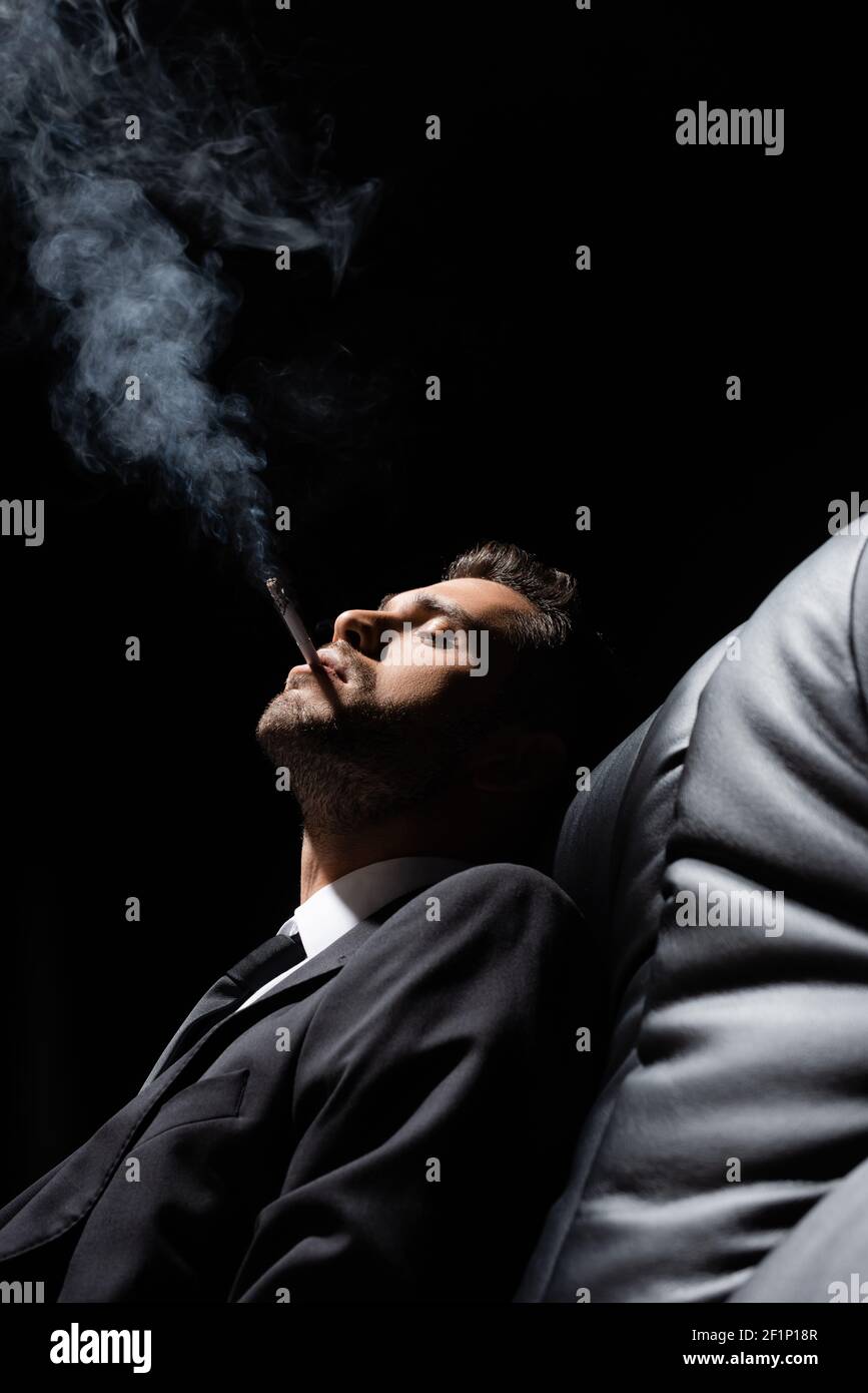 Young man in suit smoking cigarette on couch on blurred foreground isolated  on black Stock Photo