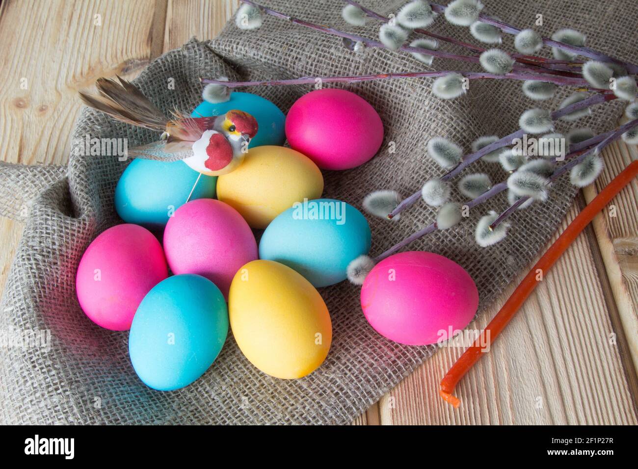 Bright colored Easter eggs lie on a natural wooden background, along with a toy bird and willow branches . Stock Photo