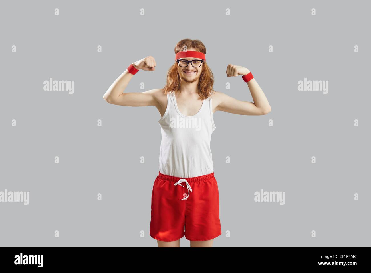 Funny skinny man in tank top and shorts showing weak muscles isolated on gray background Stock Photo