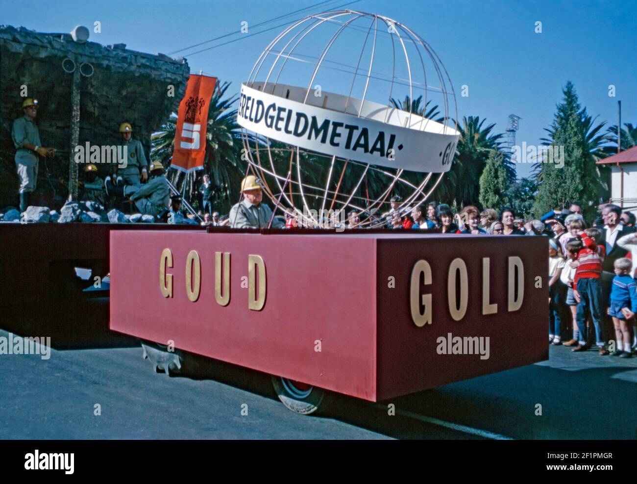 Parade float promoting South African gold mining (goud in Afrikaans), during the apartheid era, Durban, South Africa, 1966.  The tractor is pulling a trailer with gold-miners on it ‘drilling’ for the precious metal. It’s very much a white audience viewing this event. This image is from an old amateur 35mm Kodak colour transparency. Stock Photo