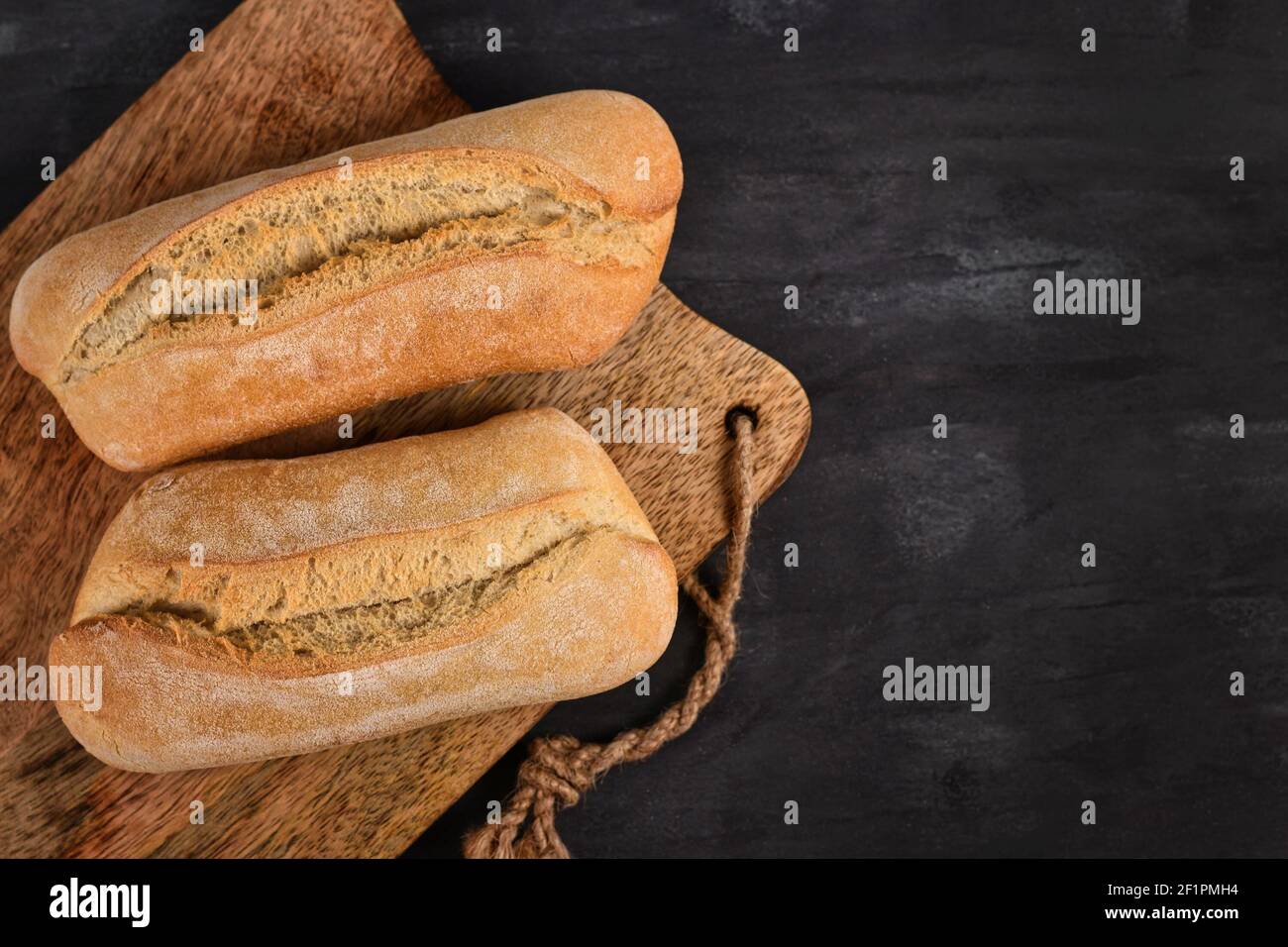 Two whole rustic bread rolls on wooden cutting board on dark table Stock Photo