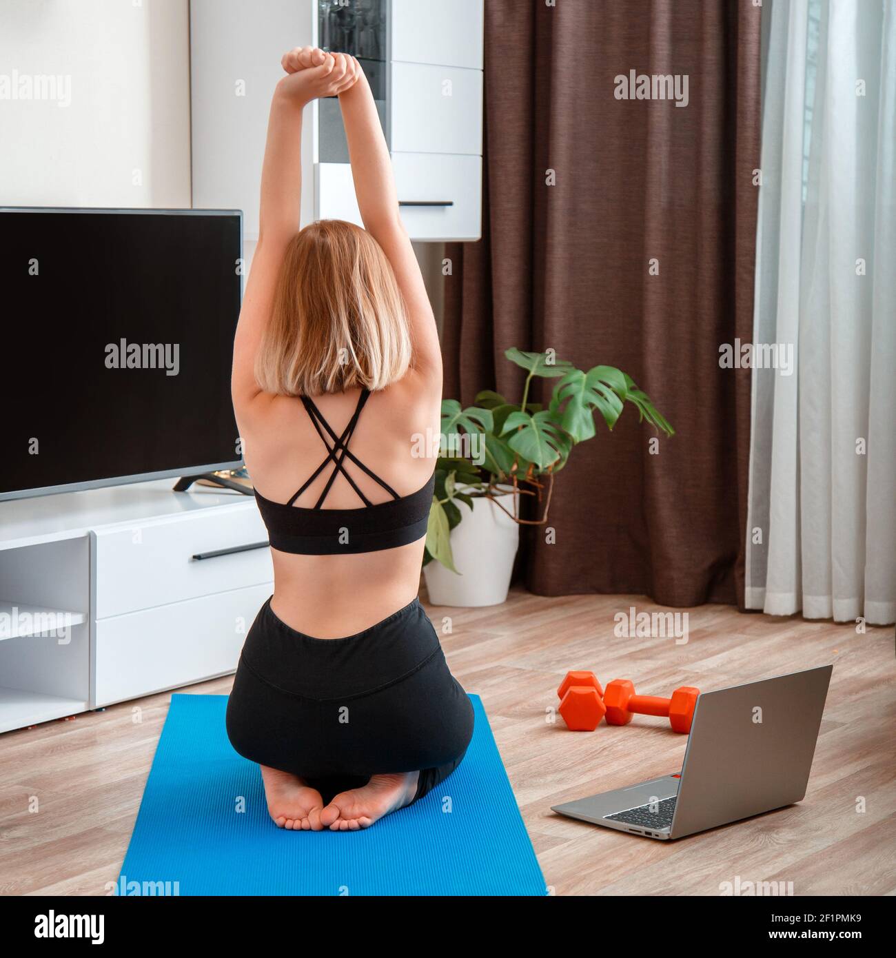 Sporty woman making fitness yoga at home via laptop by remote video call online. Young woman losing weight by online remote gym workout. Back view unr Stock Photo