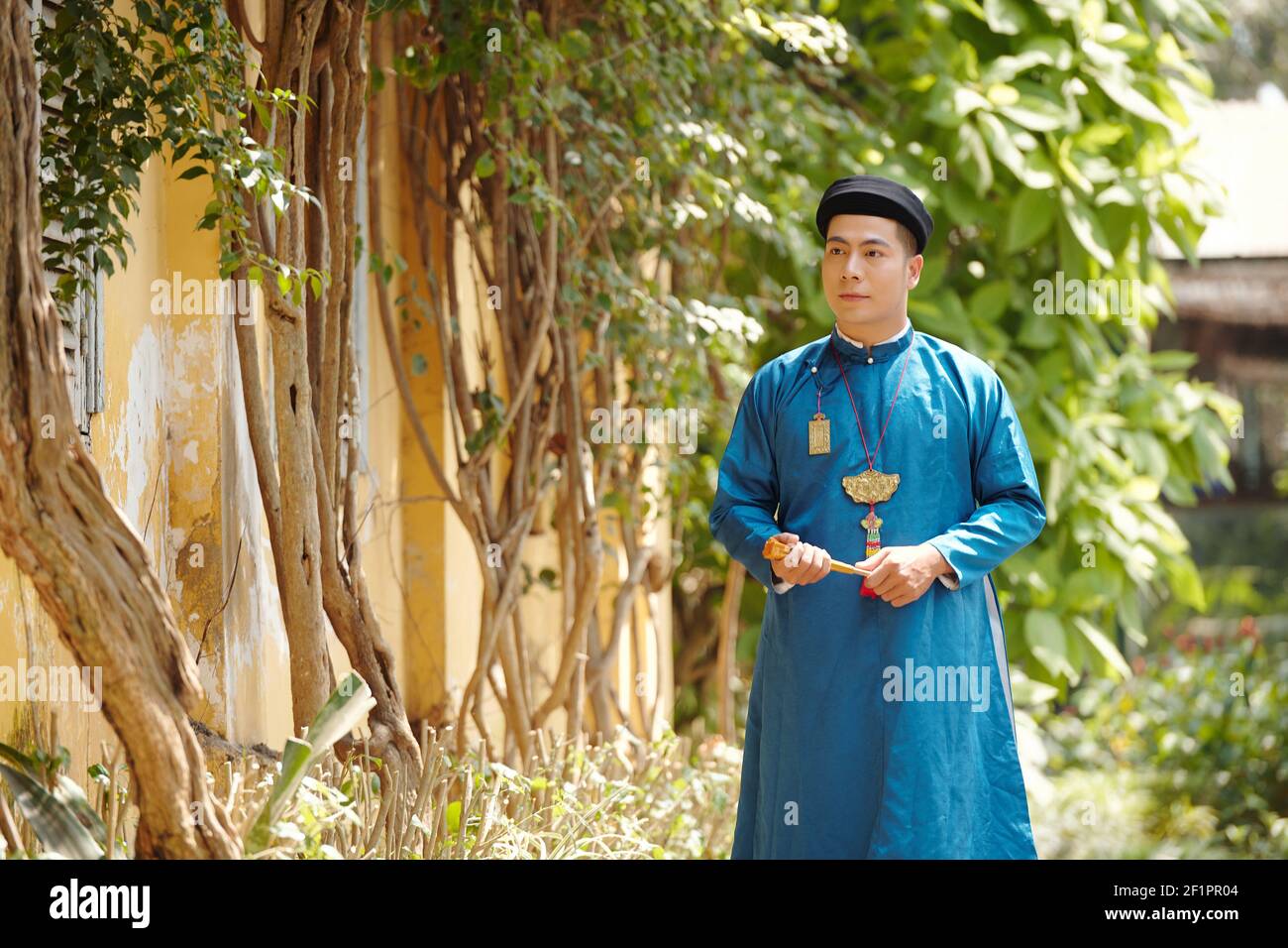 Handsome young Vietnamese man in ao dai dress walking on street with paper fan in hands Stock Photo