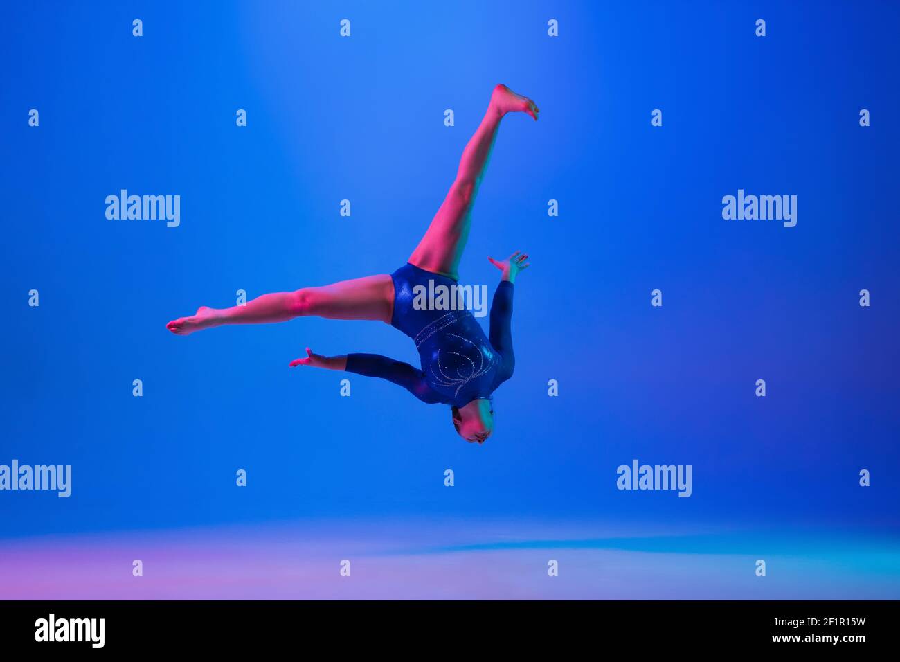 Jumping high. Young flexible girl isolated on blue studio background in neon light. Young female model practicing artistic gymnastics. Exercises for flexibility, balance. Grace in motion, sport, action. Stock Photo