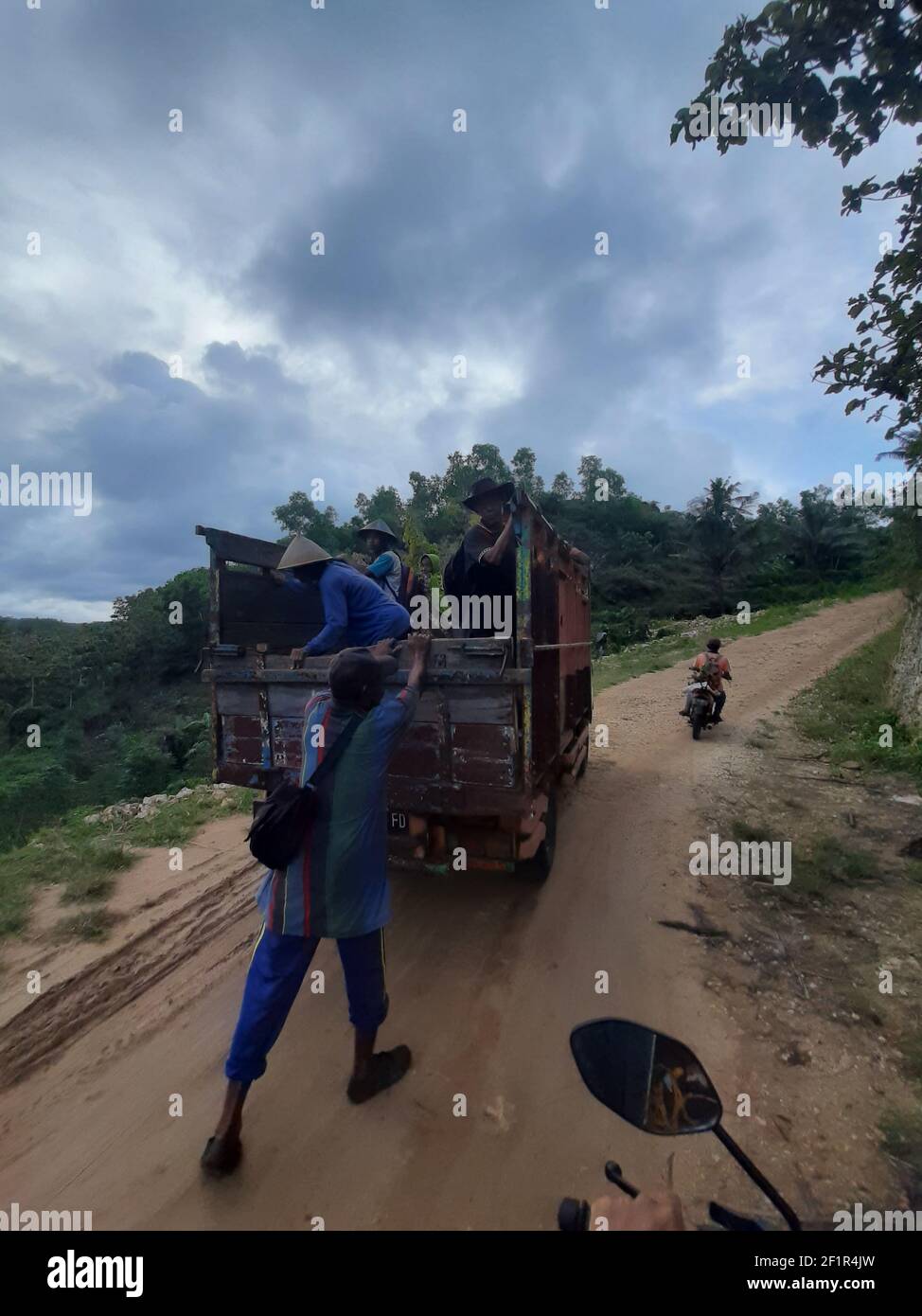GUNUNG KIDU, INDONESIA - Jun 14, 2020: POV in over head shot from a motorbike overtake a truck with a man climbing on the loading area with more peopl Stock Photo