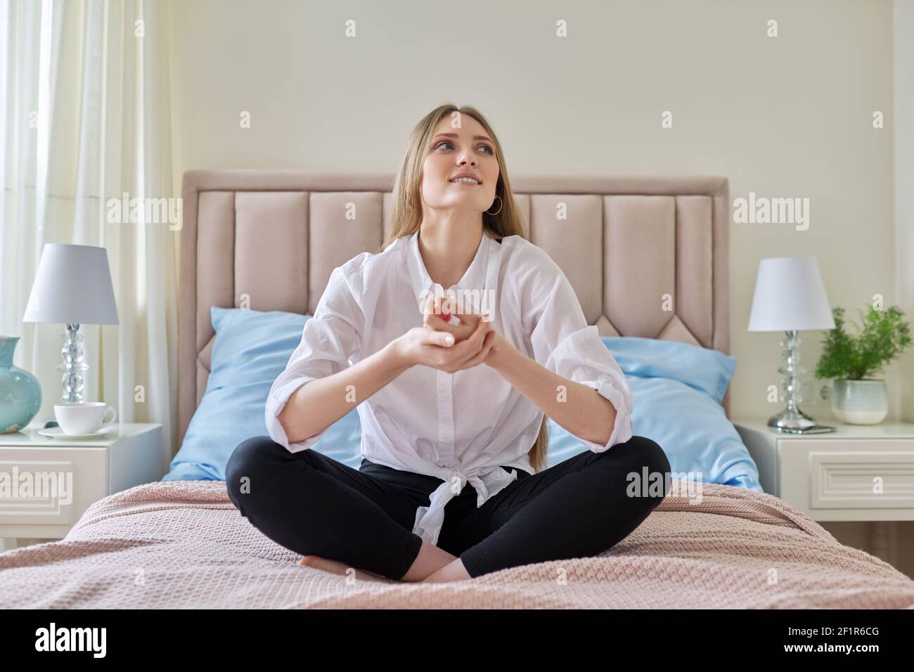 Calm young beautiful woman sitting on pastel bed in lotus position ...