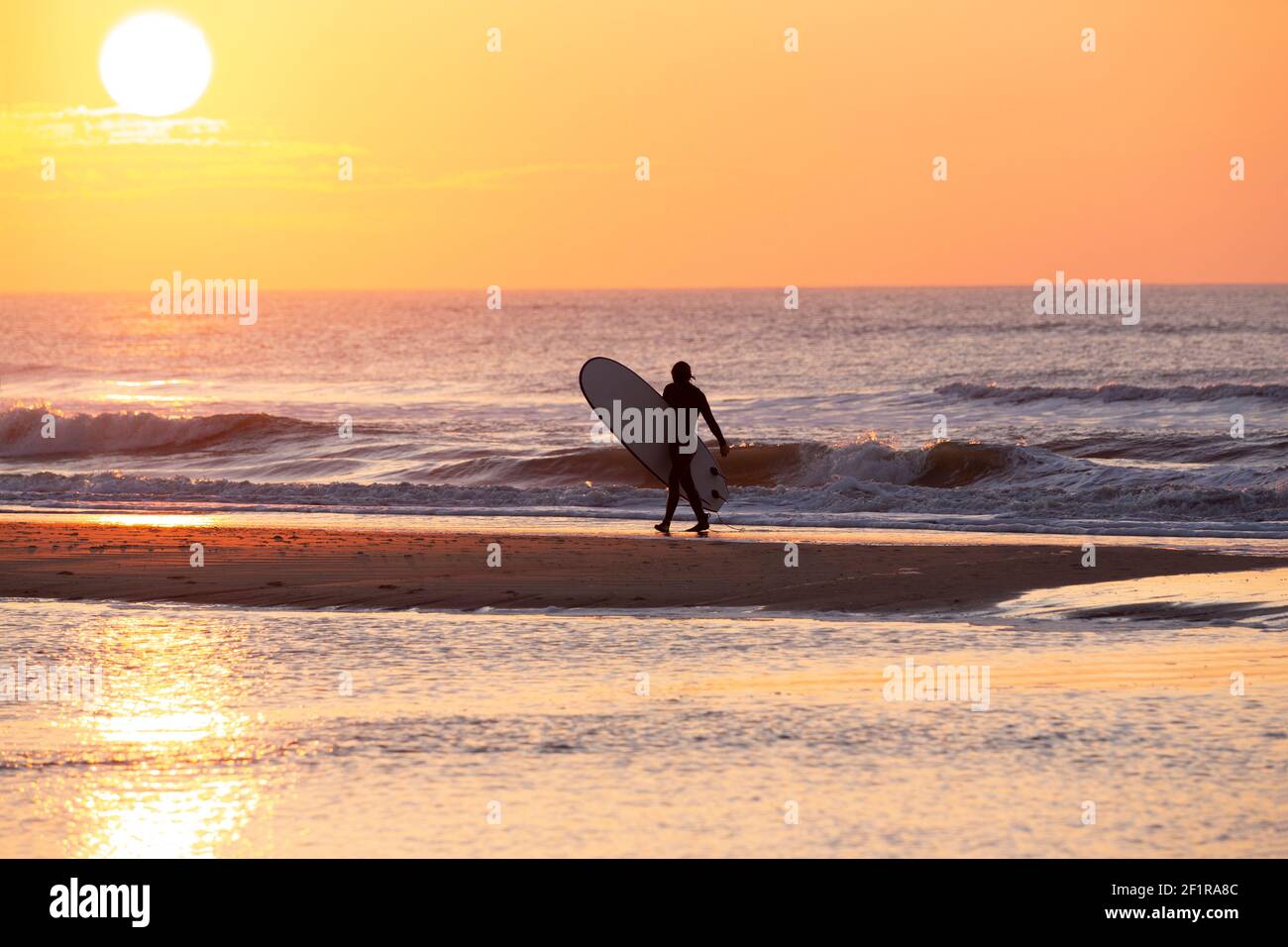 A surfer walking along the beach at sunset Stock Photo
