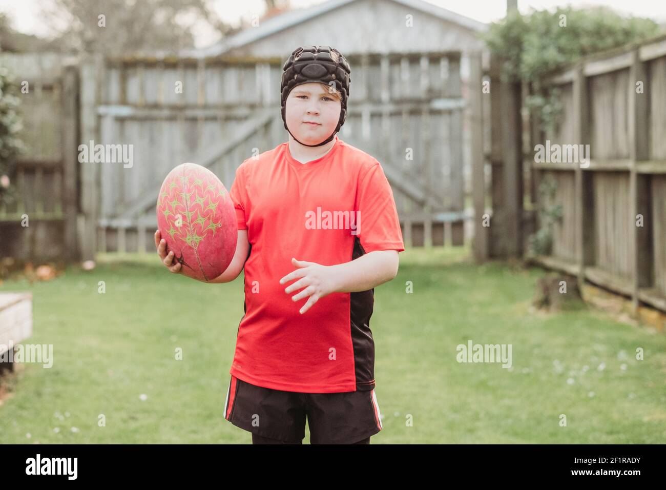 Boy wearing rugby head gear and holding rugby ball in backyard Stock Photo