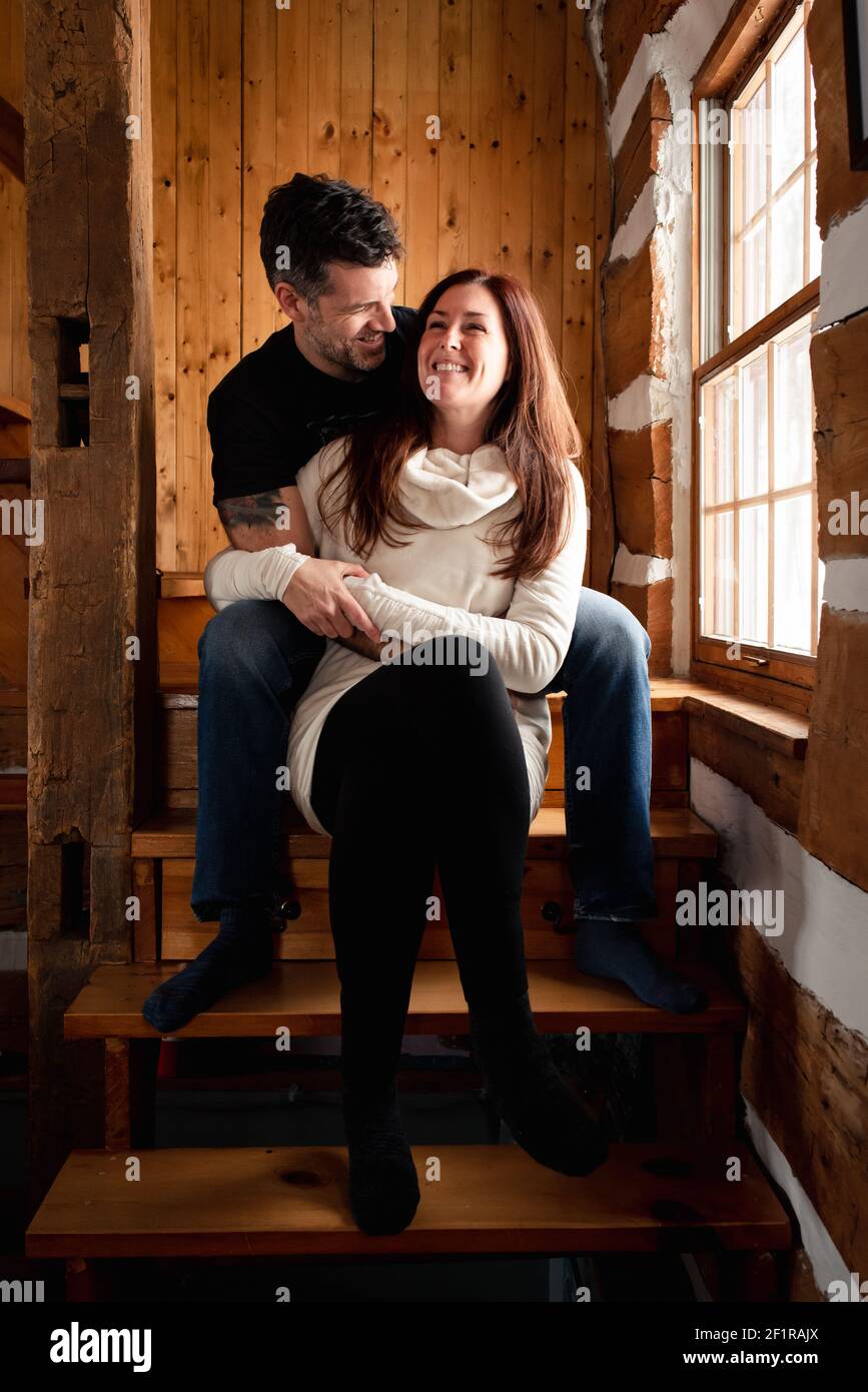 Happy couple sitting on stairs together inside rustic log cabin. Stock Photo