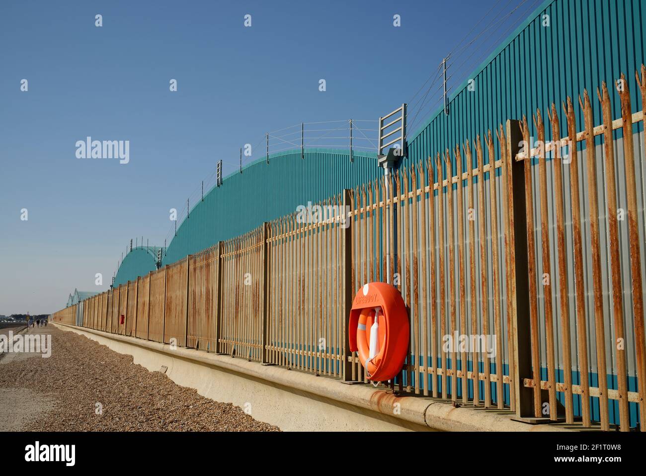 Rusty fence along a beach with a lifebuoy and turquoise industrial sheds behind. Stock Photo