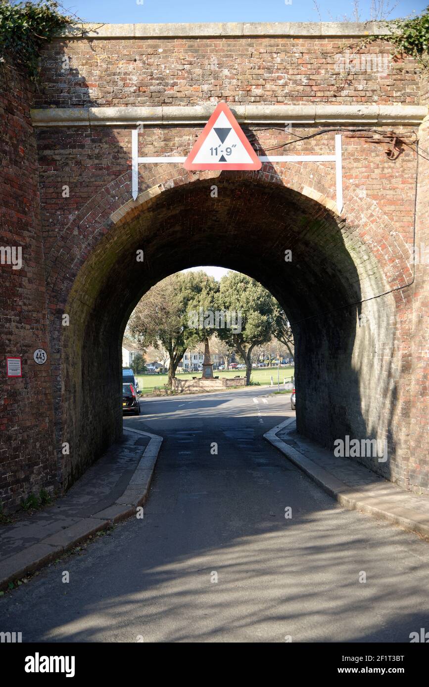 Railway bridge over a road in England. Triangular warning road sign depicts maximum height. Brick arch with a green park on the far side. Stock Photo