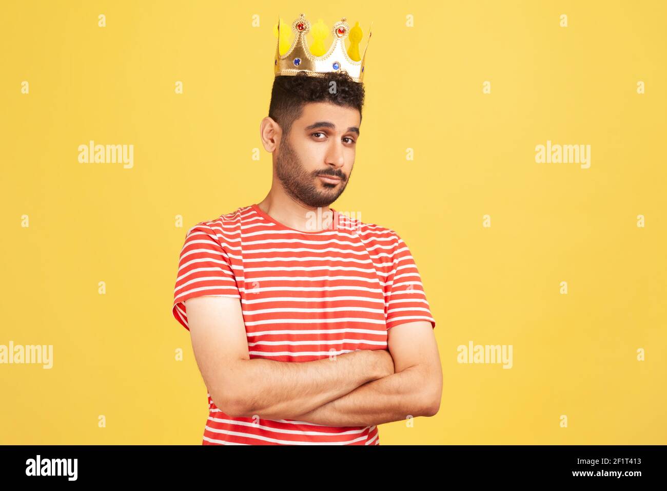 Arrogant selfish bearded man in striped t-shirt egoistically looking at camera, posing with crown on head, pretending to be king, privileged status. I Stock Photo