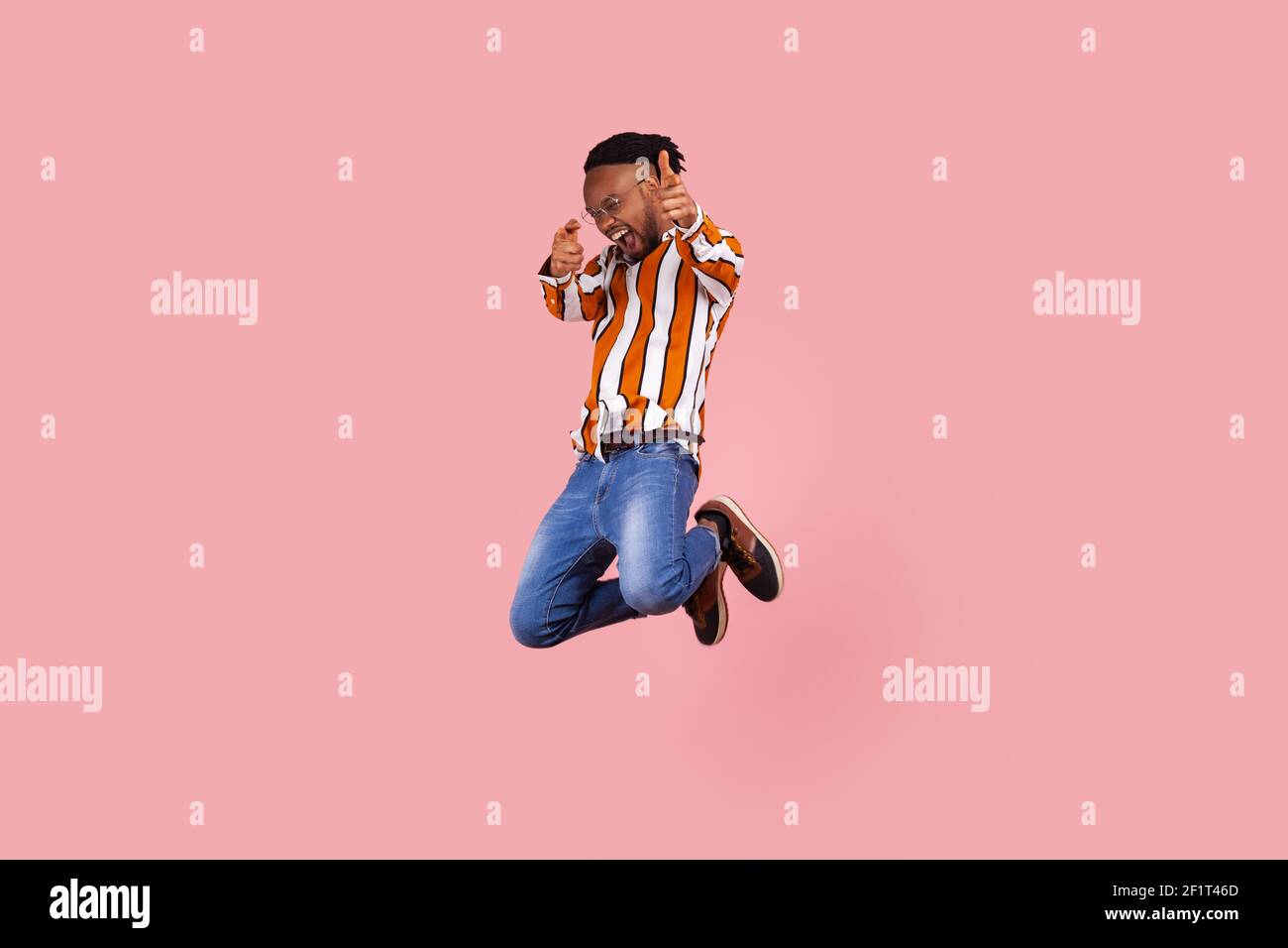 Full length happy successful african man with dreadlocks in striped shirt denim pants actively jumping pointing fingers at camera, celebrating victory Stock Photo