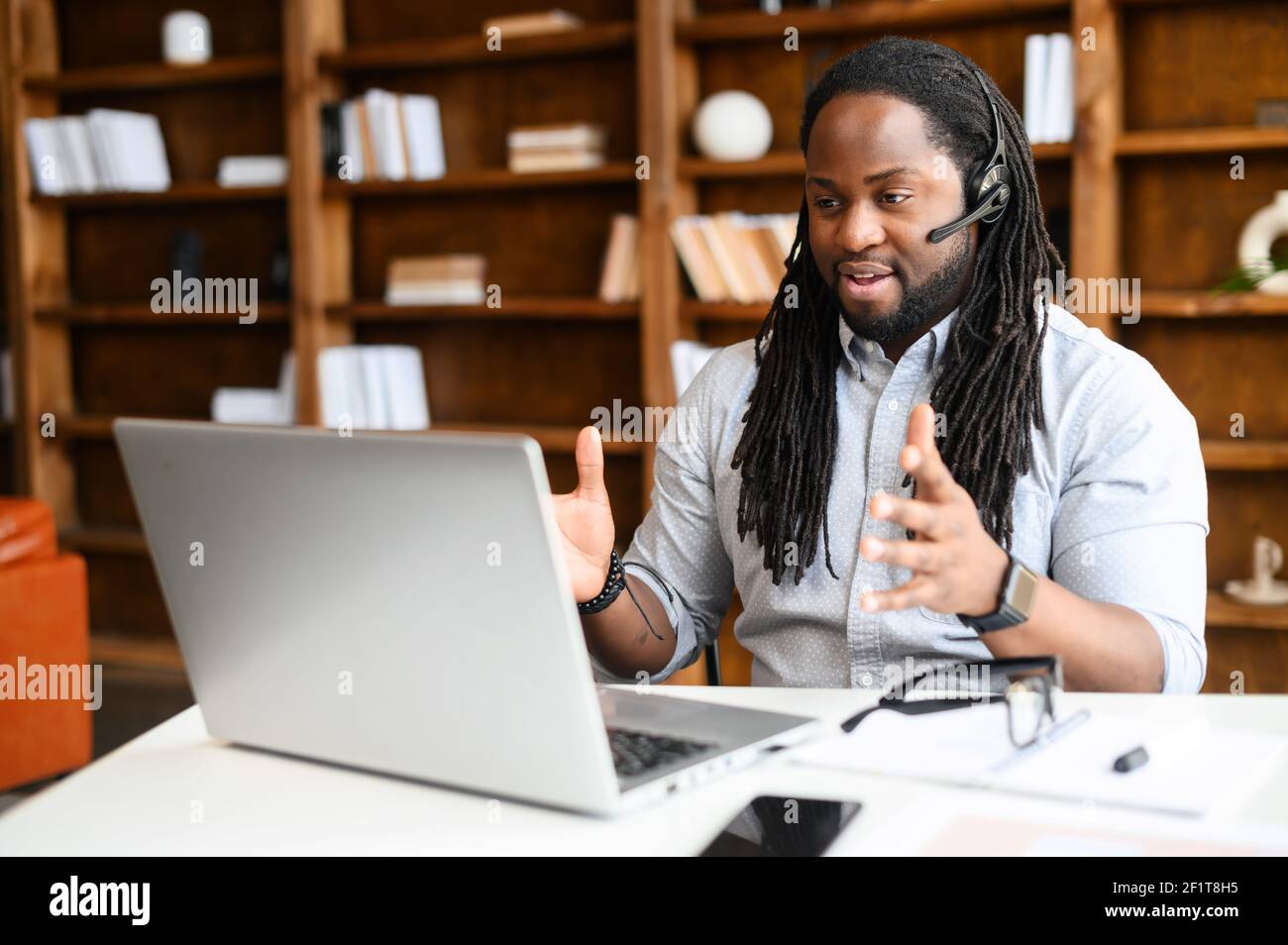 Diverse office employees concept. A focused African-American man with a dreadlocks  wearing wireless headset is using laptop for online video conversation,  talking with customers or coworkers online Stock Photo - Alamy