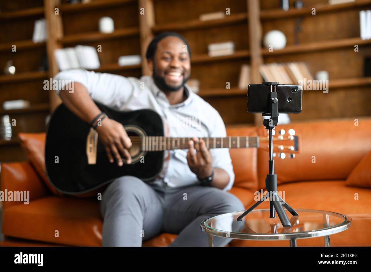 Selective focus at the smartphone on the tripod in front of African guy playing guitar, a vlogger making a content, records himself on the smartphone, performing online Stock Photo