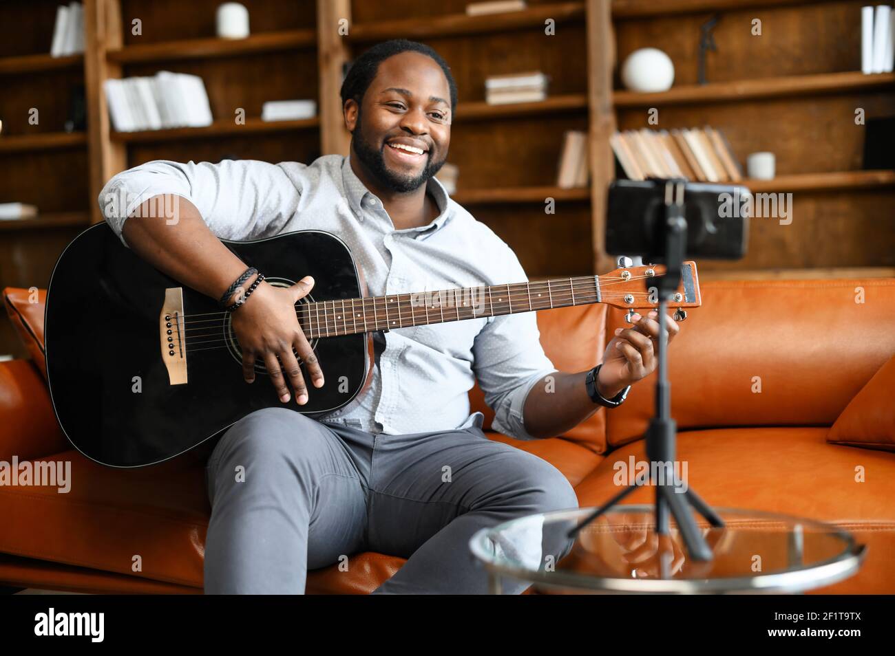 The African-American guy with a guitar is performing online, a biracial smiling man sits on the sofa, play acoustic guitar and streaming online, recording himself on the smartphone, holding a webinar Stock Photo