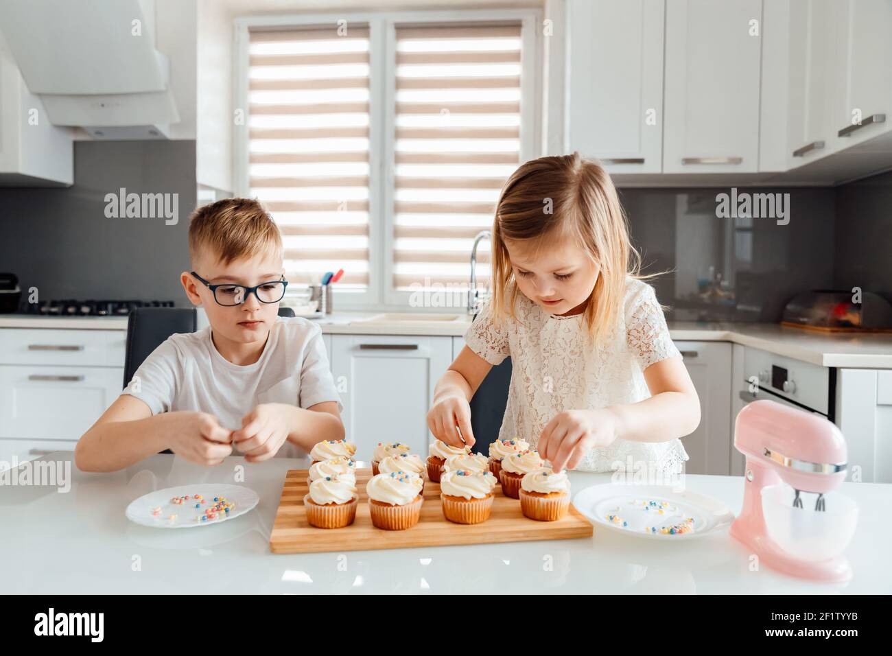 Two children decorate cupcakes by sprinkling icing balls on them Stock Photo