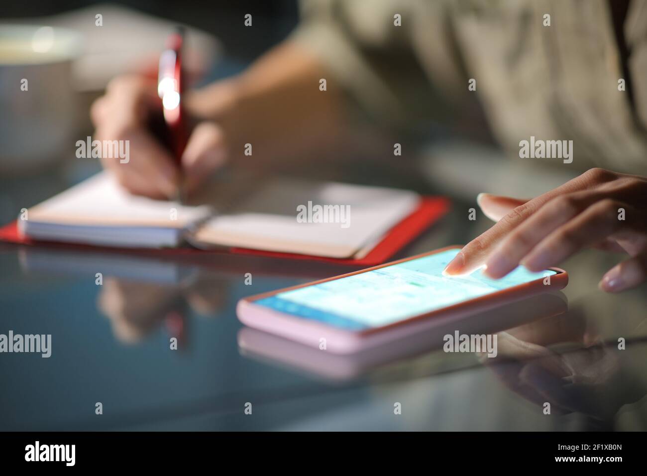 Close up of a woman hands checking smart phone calendar and writing on agenda in the night at home Stock Photo