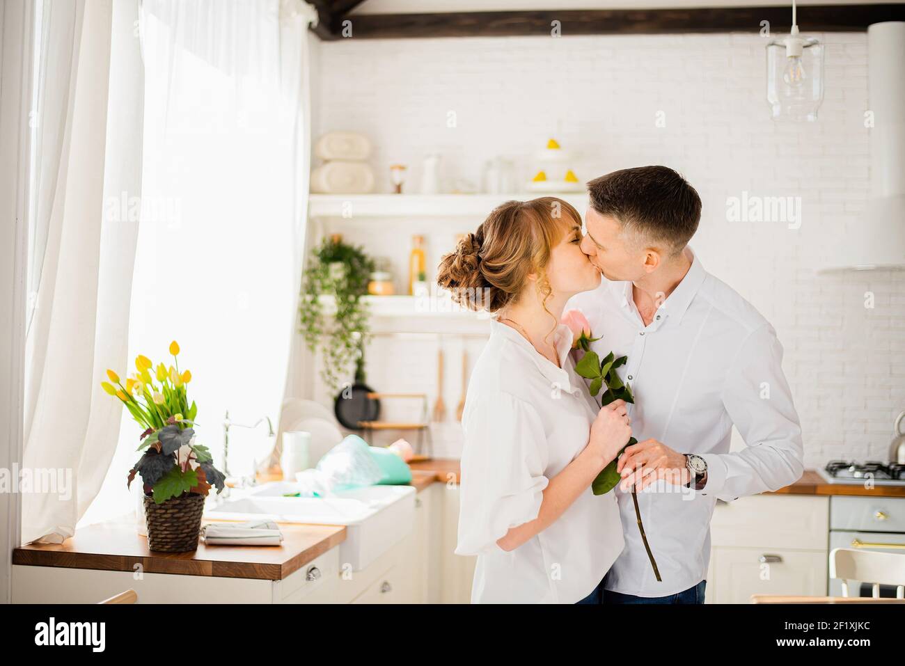 Kiss of a couple in love in the kitchen with a rose in their hands Stock Photo