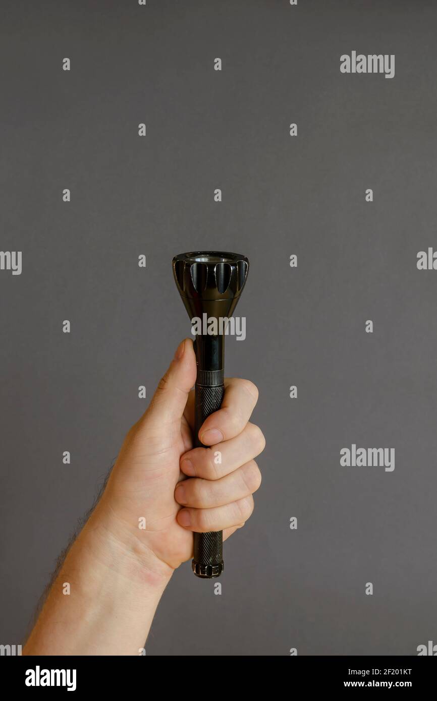 A hand holds a black modern LED flashlight on a gray background. Male hand and a metal flashlight. Side view. Selective focus. Stock Photo