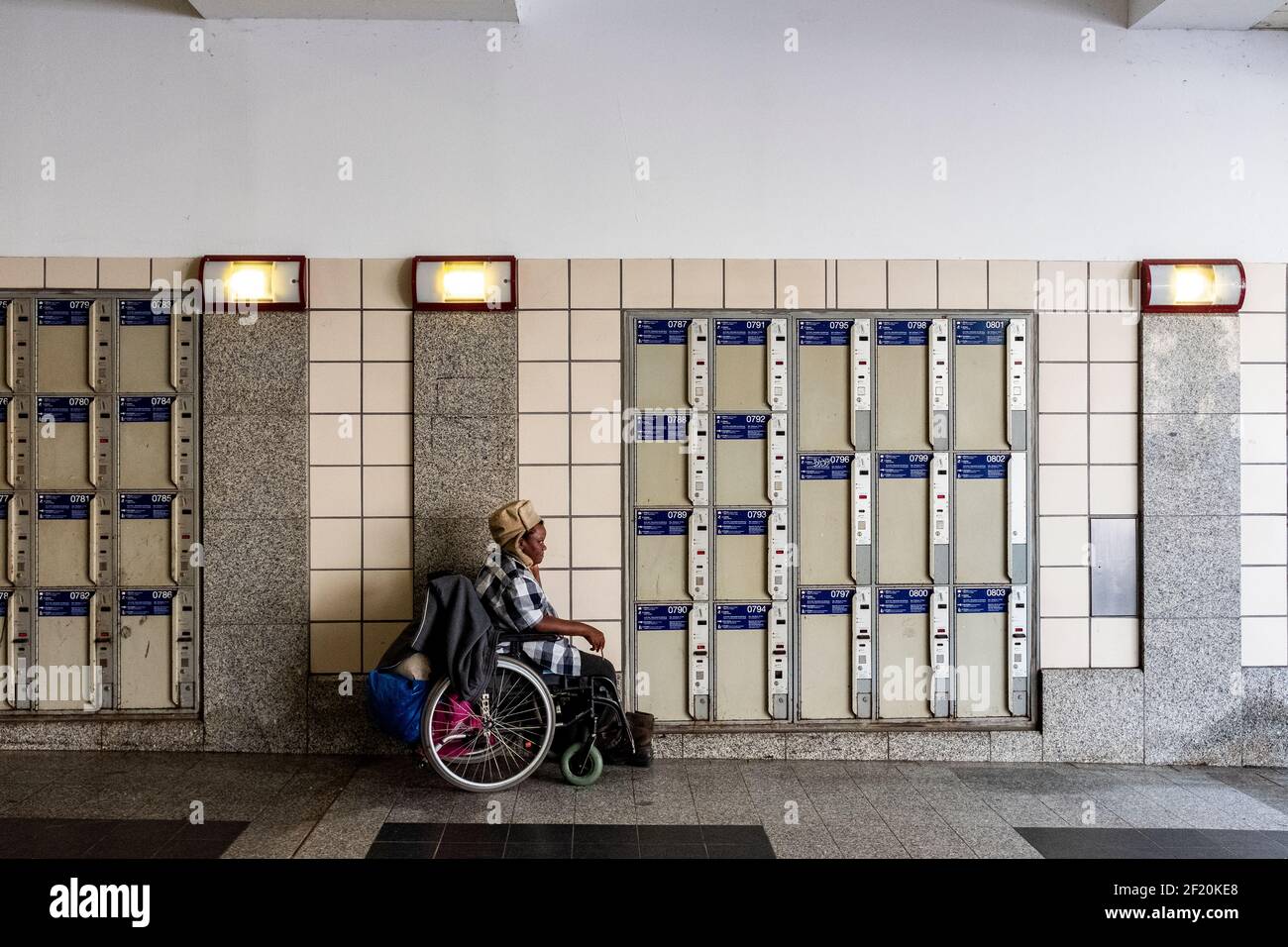 Berlin, Germany. Colored homeless woman, killing time and passing her day, sitting in her wheelchair, parked in front of the Bahnhof Zoo Luggage Lockers. Stock Photo