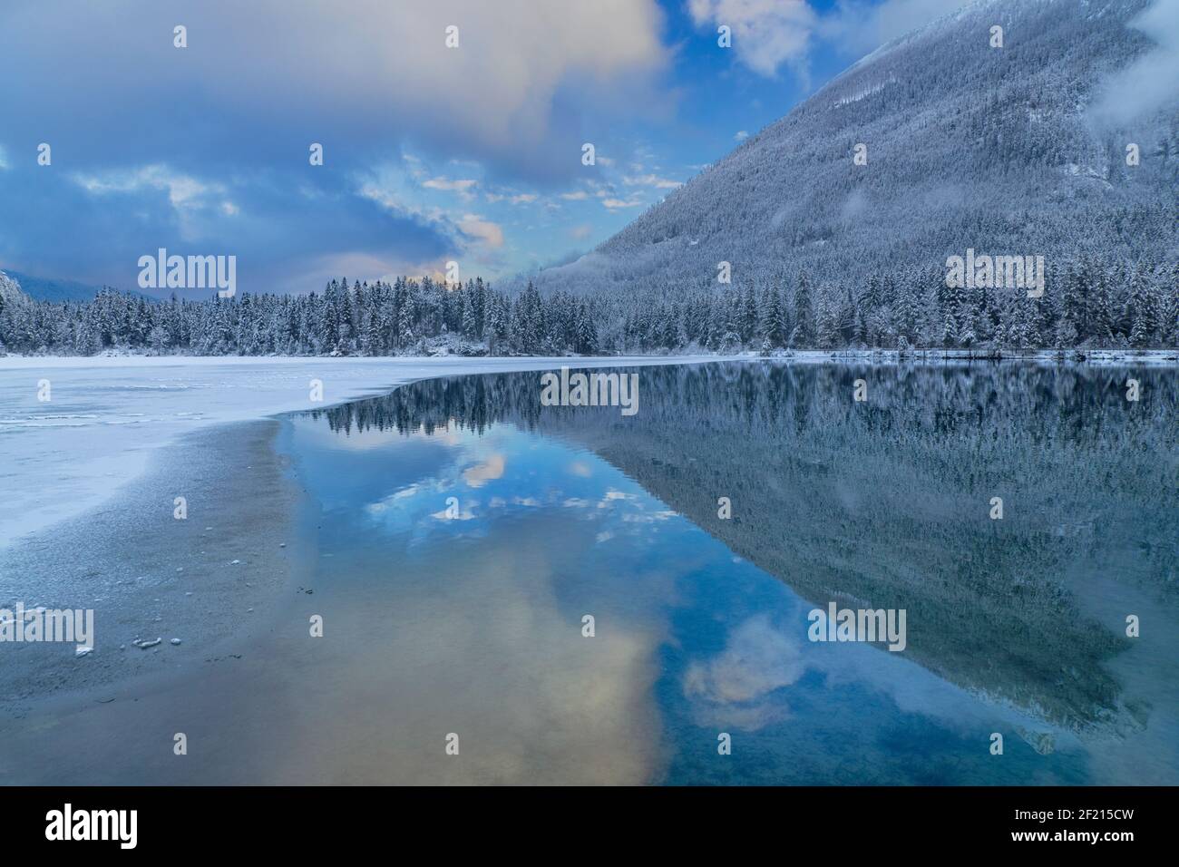 Germany, Bavaria, Berchtesgaden, Berchtesgadener Alps, Partially snow covered and frozen Lake Hintersee with reflection of sky, hillside and clouds in the unfrozen section. Stock Photo