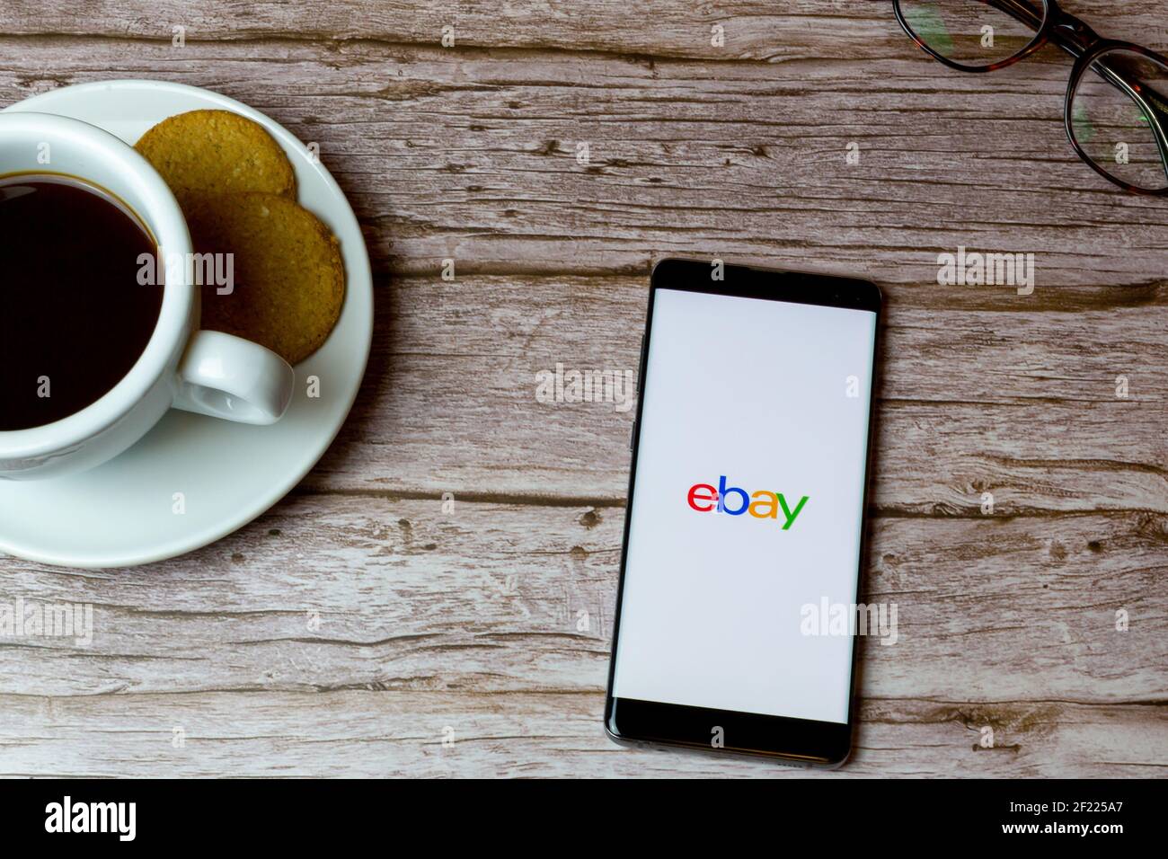 A Mobile phone or cell phone laid on a wooden table with an Ebay app opening also a coffee and glasses Stock Photo
