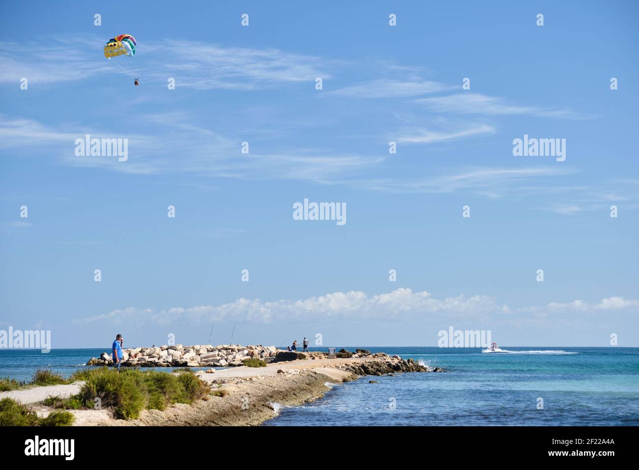 A couple enjoying a tandem parasailing ride hand in the air high above anglers fishing on the causeway entrance to the Grand Canal, Playa de Muro, Mal Stock Photo