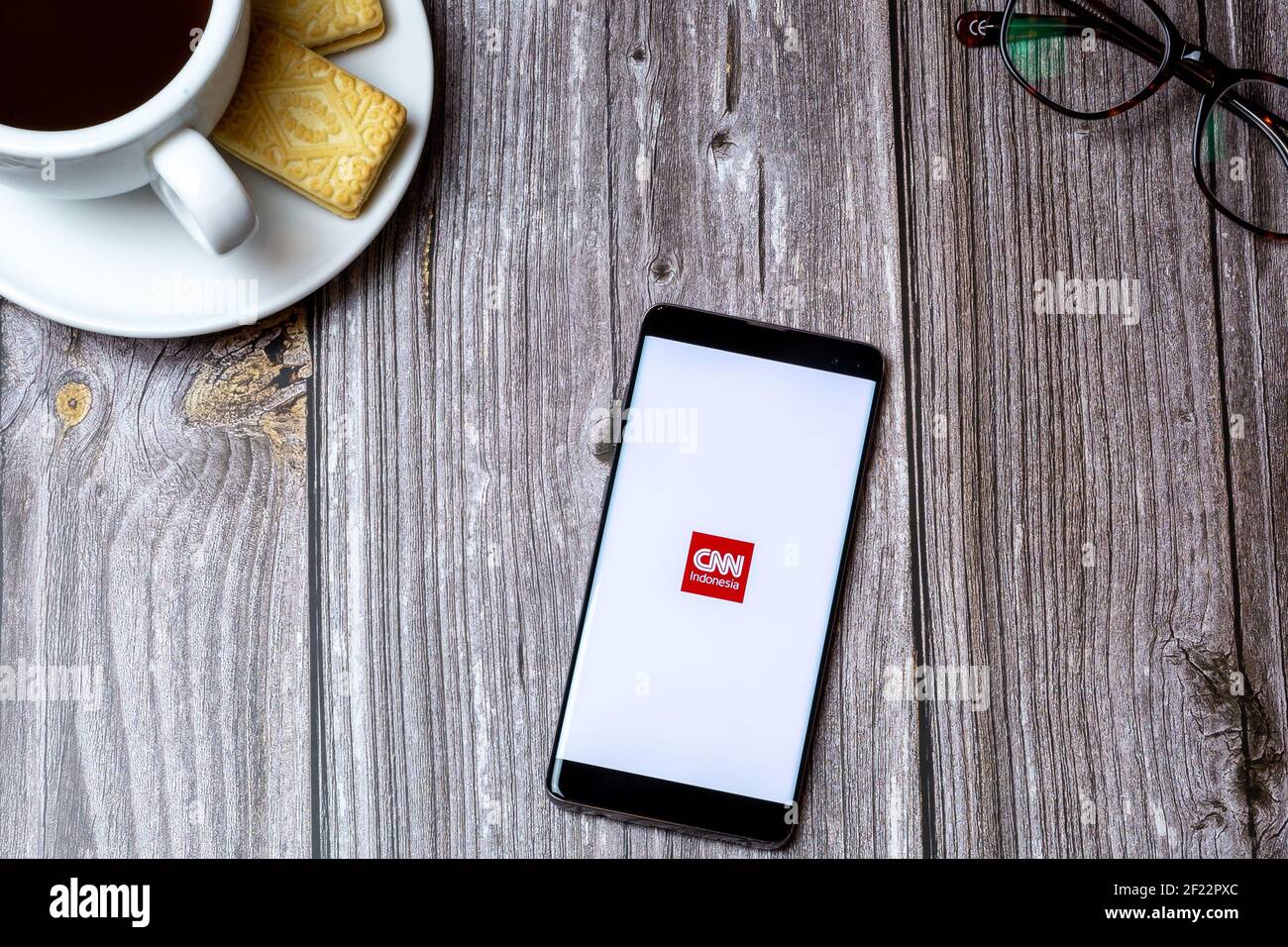 A mobile phone or cell phone on a wooden table with the CNN Indonesia app open next to a coffee and glasses Stock Photo