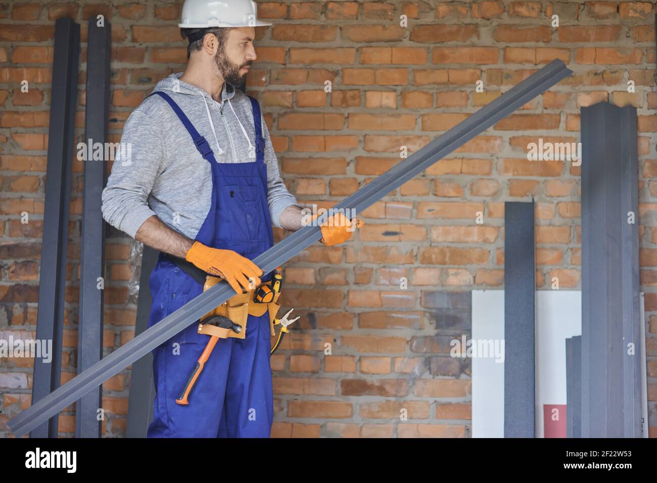 Confident male builder wearing overalls and hard hat looking focused while holding a metal stud for drywall, working on house construction. Building house, profession, safety concept Stock Photo