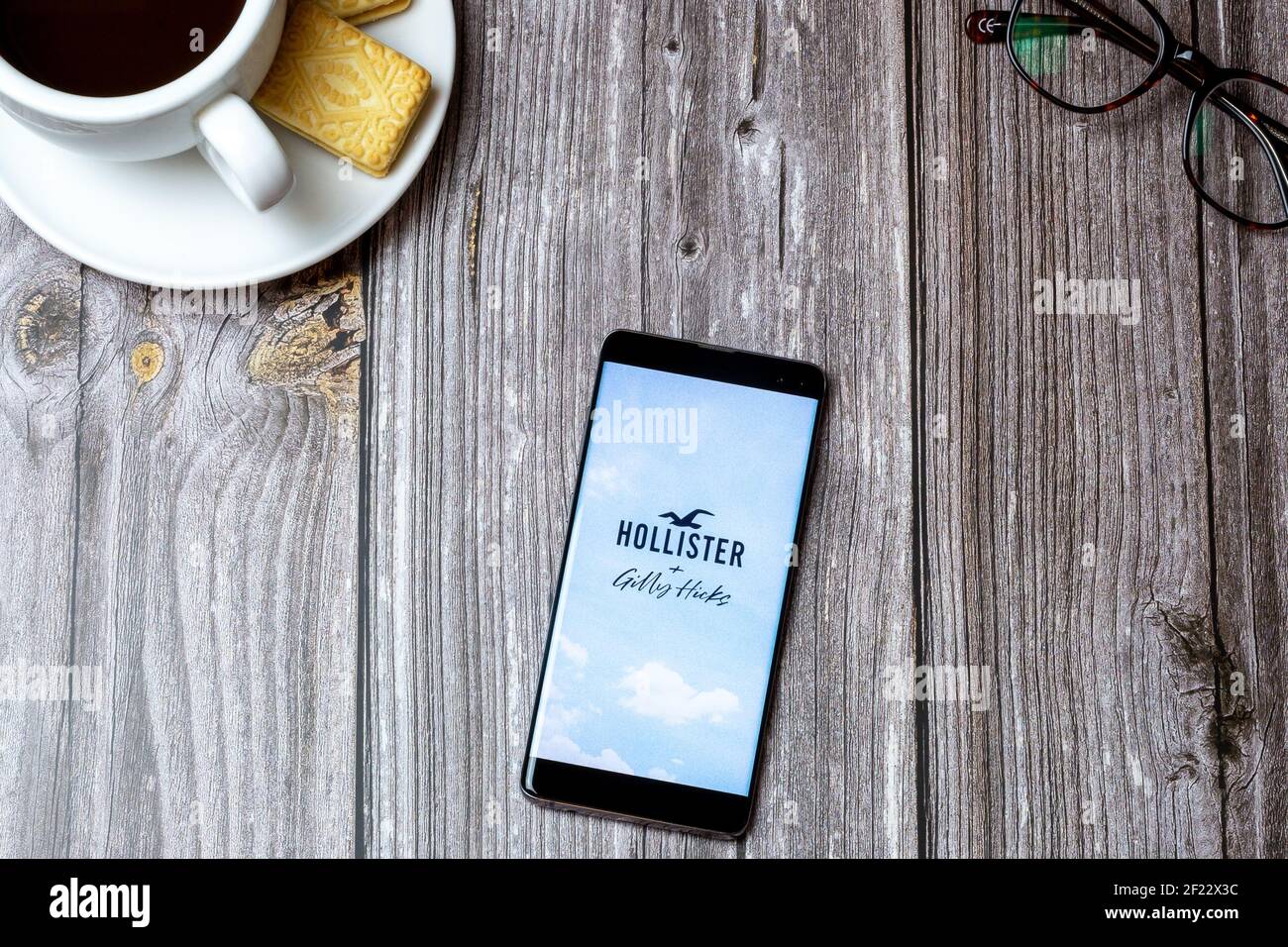 A mobile phone or cell phone on a wooden table with the Hollister app open next to a coffee and glasses Stock Photo