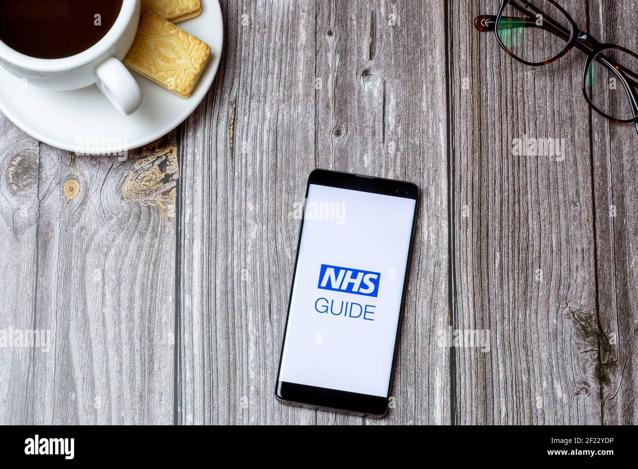 02-24-2021 Portsmouth, Hampshire, UK A mobile phone or cell phone on a wooden table with the NHS Guide app open next to a coffee and glasses Stock Photo