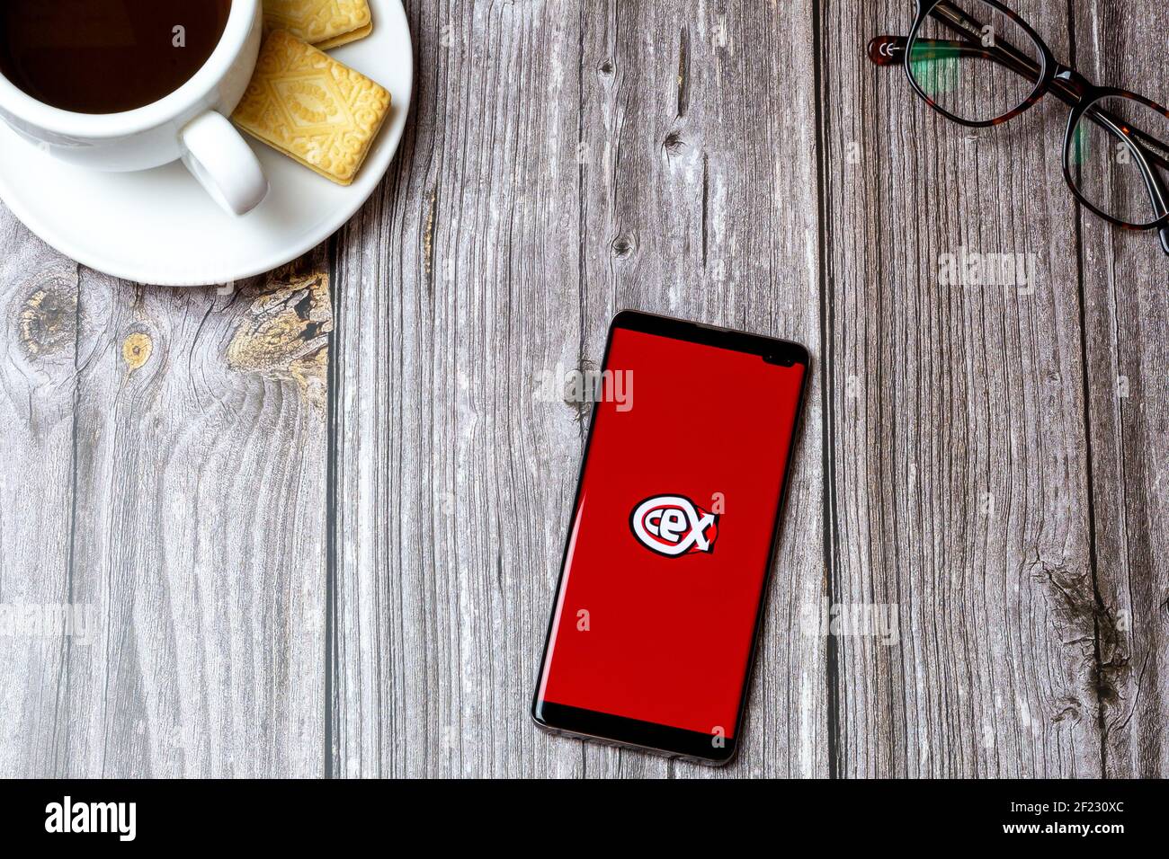 A mobile phone or cell phone on a wooden table with the CEX app open next to a coffee and glasses Stock Photo