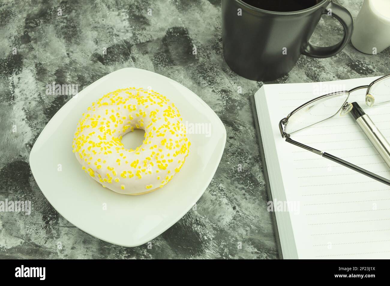 Round donut in chocolate on a white plate. Donut covered with white chocolate and sprinkled with lemon zest. Donut and coffee on the table. Space for Stock Photo