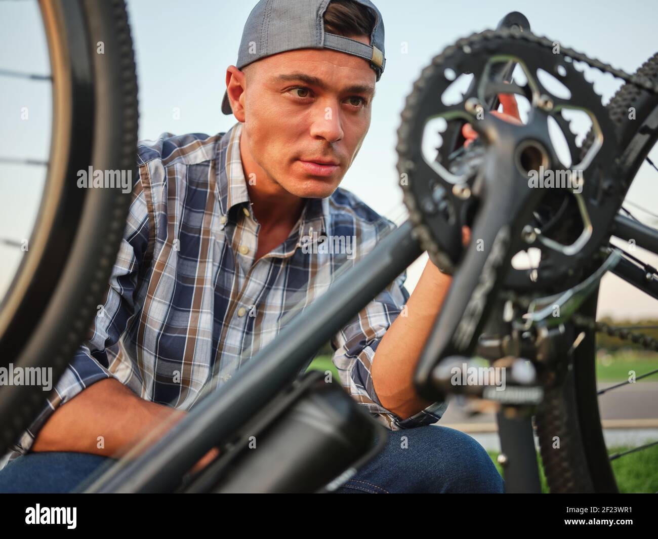 Portrait of handsome caucasian man looking concentrated while fixing pedals on his bike, kneeling on grass. Repairing, transportration concept Stock Photo