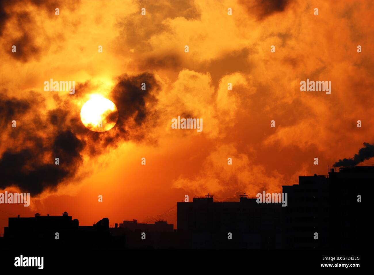 Sunrise on colorful dramatic sky, orange sun shining through dark clouds above the buildings silhouettes. Picturesque city landscape for horror Stock Photo