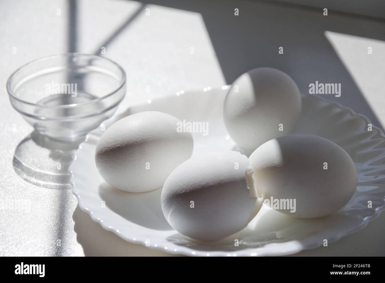 four boiled eggs in elegant little white porcelain plate. One is slice open. on clean windowsill in the sunlight Stock Photo