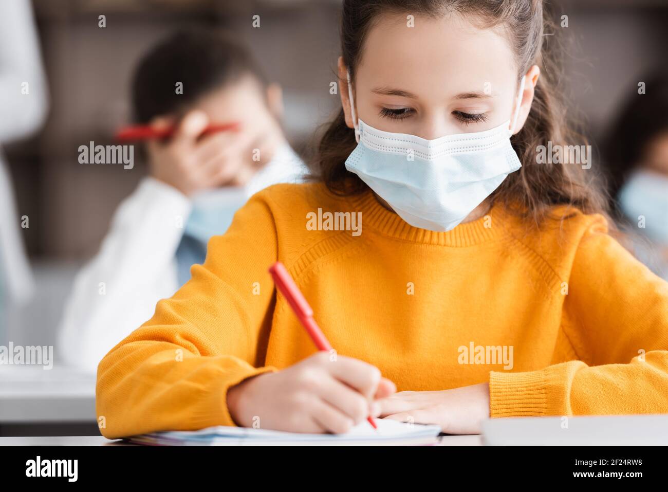 schoolgirl in medical mask writing in notebook during school lesson Stock Photo