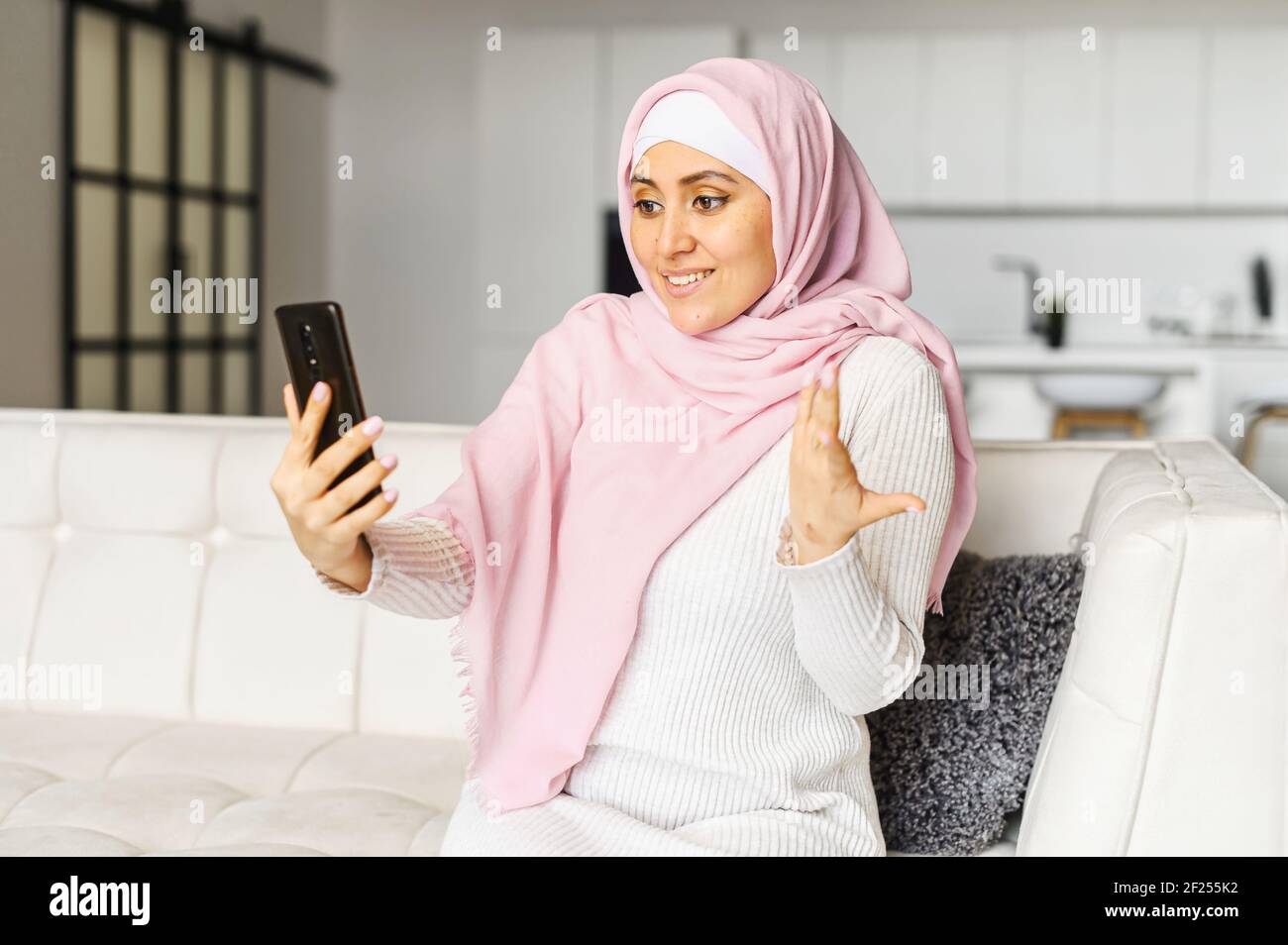 Young Muslim Arabic mixed-race woman talking online on mobile phone, sitting on the couch in studio apartment, video-calling with family coworkers, chatting, gesturing, discussing news with a friend Stock Photo