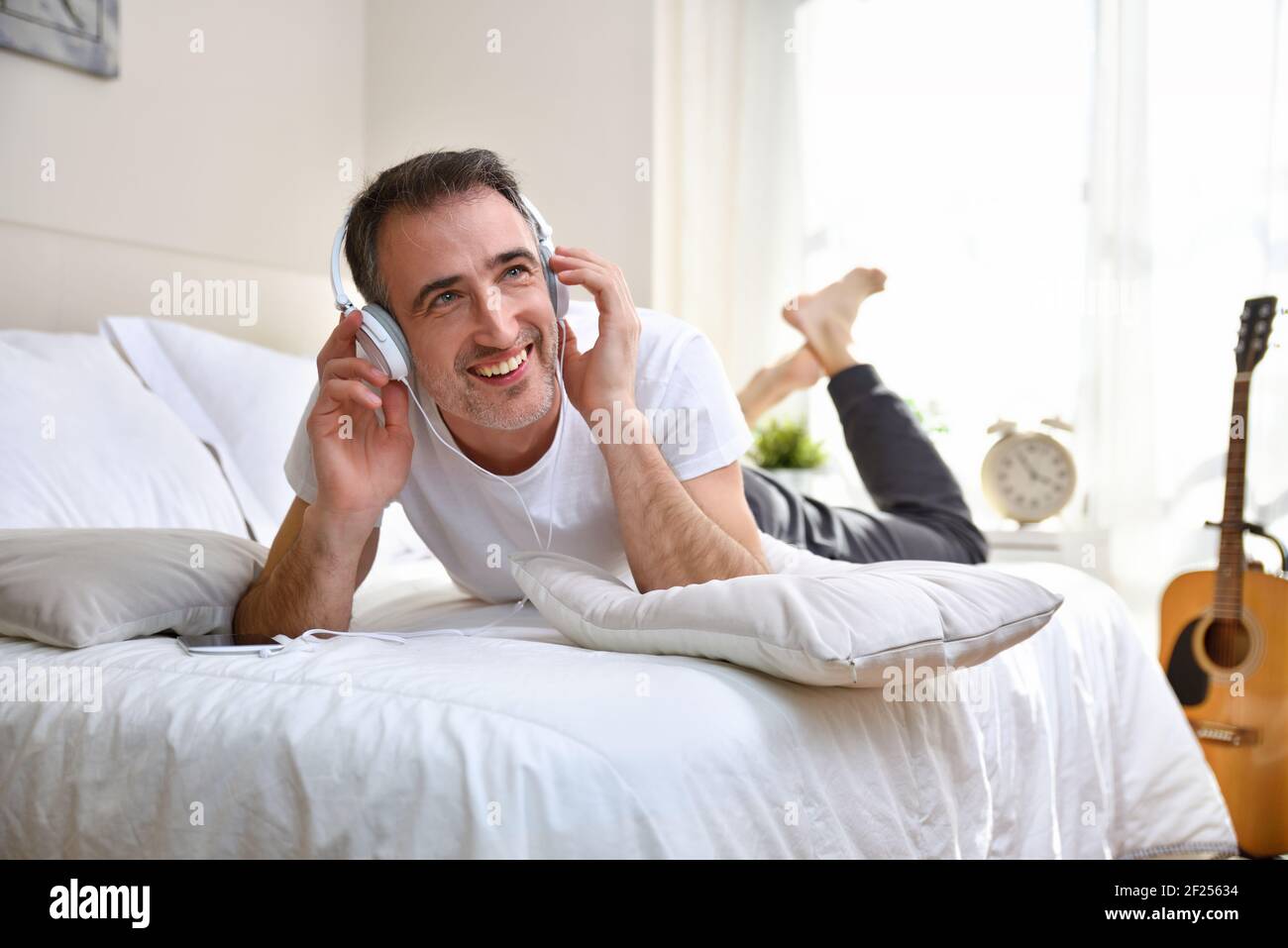 Happy adult man smiling listening to music lying in bed with white ...