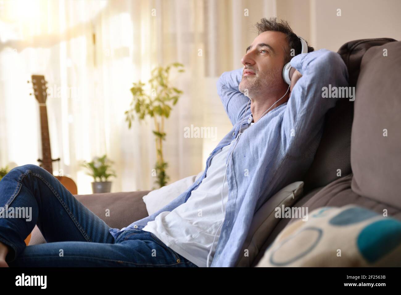 relaxed man placidly listening to music from white headphones staring blankly sitting on the couch at home Stock Photo