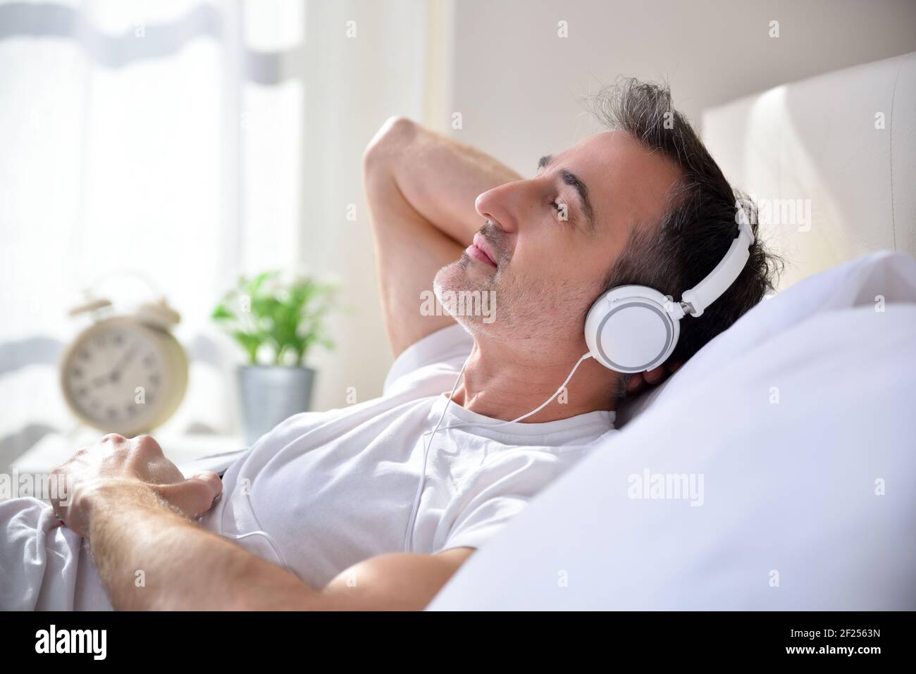 Happy and relaxed adult man with closed eyes listening to music lying in bed with window in the background entering the morning light. Stock Photo