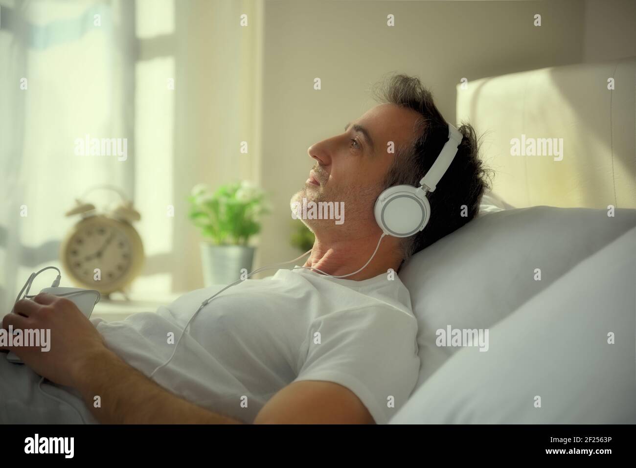 Melancholic adult man with lost eyes listening to music with white headphones from the mobile lying on the bed entering sunset light Stock Photo