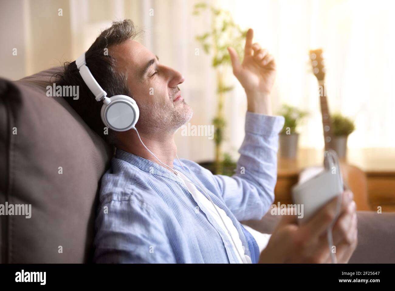 Detail of man sitting on the sofa at home listening to music placidly from a mobile phone with white headphones entering a warm sunlight through the w Stock Photo