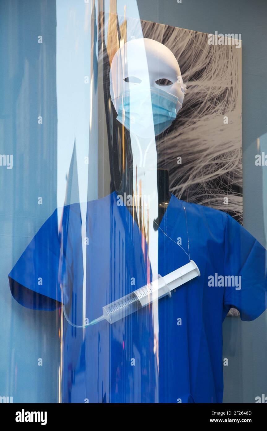 Sinister masked mannequin clothed in blue surgical scrubs, with a syringe. Window display of a hairdressers salon, during the Coronavirus pandemic. UK. Stock Photo