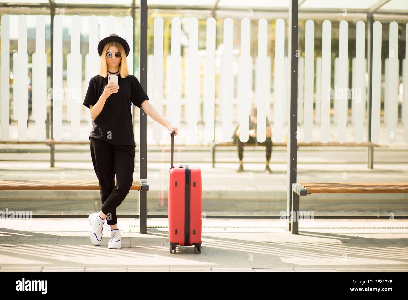 Traveler tourist woman in black casual clothes, sunglasses and hat with red suitcase use mobile phone to buy tickets online and sit on bus station. Female passenger travel abroad on weekends getaway. Air flight journey concept Stock Photo