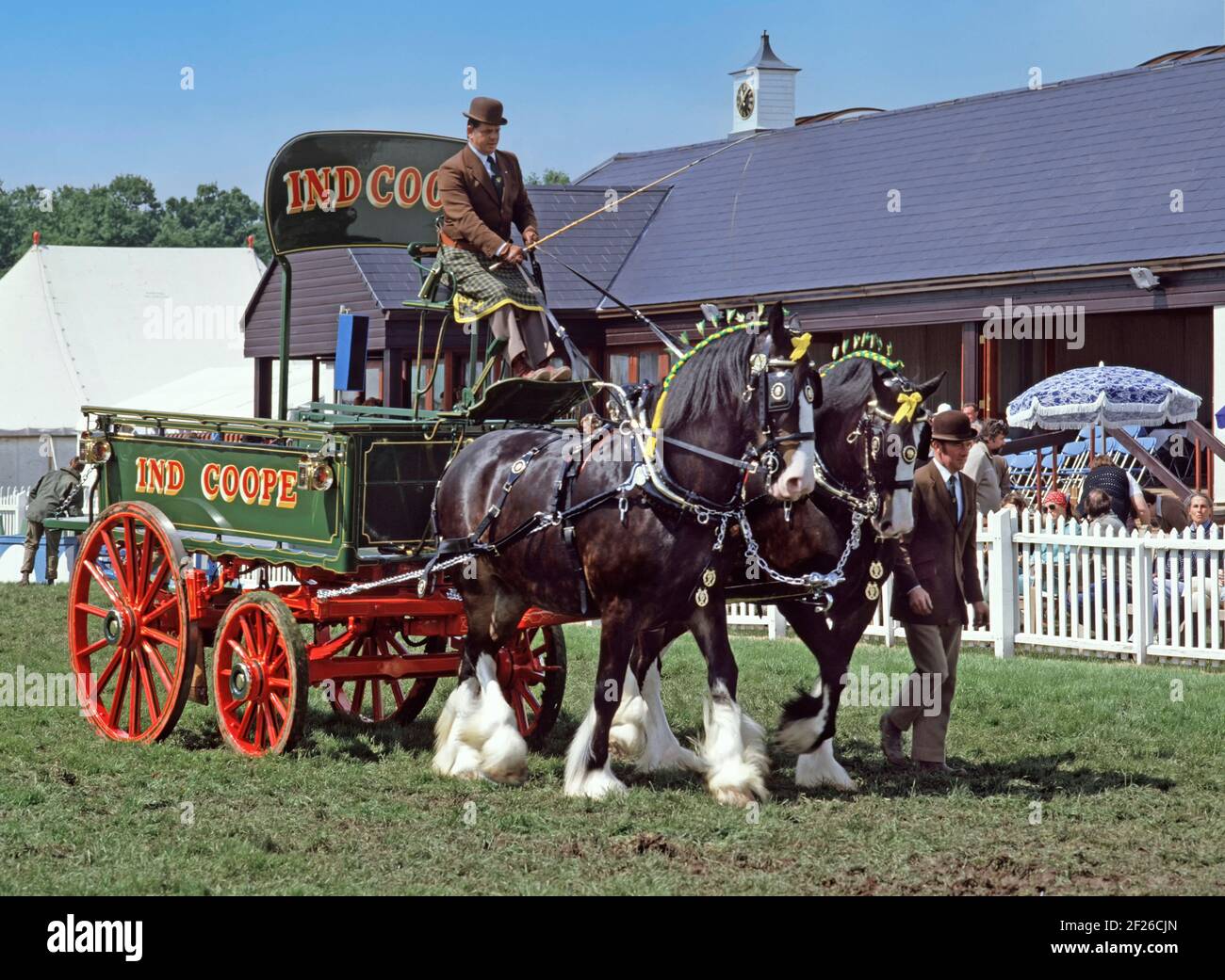 Historical 1985 archive view of Ind Coope brewery dray being pulled by two heavy Draught Shire horses with decorated mane ribbons driver and mate in company uniform parading and promoting the business brand in the main arena at a 1900s Essex County agriculture show event at Great Leighs showground on a blue sky sunny summer 80s day for this way we were archival image near Chelmsford Essex England UK Stock Photo